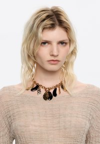 Collier en chaîne dorée avec des pendentifs noirs de différentes formes, présentant un design texturé. Coiffé d'un haut beige clair.