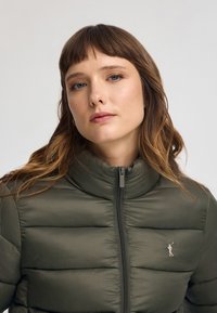 Grön puffjacka med hög krage, dragkedjestängning och horisontell sömnad. Har en liten logotyp på vänster bröst.