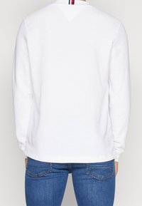 Vit sweatshirt med långa ärmar, ribbad textur och en liten triangulär detalj på nacken bak med horisontella röda och blå ränder.