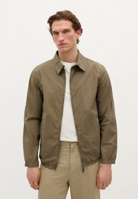 Khaki bomberjakke med lynlåslukning, skjortekrave og elastiske manchetter. Bæres over en hvid skjorte, sat sammen med lys beige bukser.