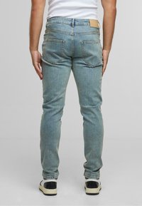 Muškarac u svijetloplavim uskim trapericama, bijeloj majici bez rukava i bijelim tenisicama s crnim petama, stoji naslonjen na jednobojnu sivu pozadinu.