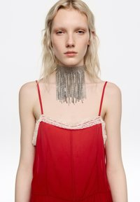 Młoda kobieta o blond włosach nosi czerwoną, bezrękawnikową sukienkę z kremową koronkową lamówką i dużym srebrnym naszyjnikiem z frędzlami w formie łańcucha na białym tle.