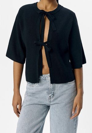 Schwarzes, kurzärmeliges Stricktop mit zwei vorderen Schnürverschlüssen, getragen zu hellblauen, hoch taillierten Jeans, wobei Bauch und Unterarme sichtbar sind.