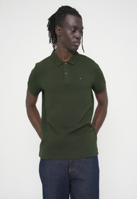 Σκούρο πράσινο πόλο μπλουζάκι από βαμβάκι, με γιακά, κοντά μανίκια και μικρό λογότυπο της μάρκας στο στήθος. Φοριέται με σκούρο τζιν.