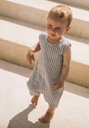 Tout-petit aux cheveux clairs et courts, portant une robe sans manches grise à carreaux, marchant pieds nus sur des marches en pierre claire sous la lumière du soleil.