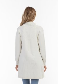 Knælængde creme cardigan med struktureret strikmønster, der har en sjal-krave og ribbede manchetter, set bagfra.