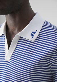 Sininen ja valkoinen raidallinen polo-paita, jossa on valkoinen kaulus ja sininen emblem kauluksessa. Puuvillakangas, jossa on V-pääntie.