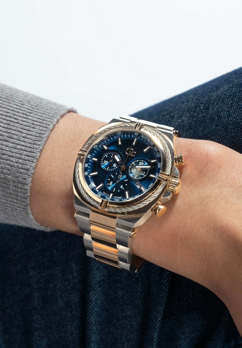 Armbanduhr mit blauem Zifferblatt, Gehäuse aus Gold und Silber, Chronographen-Subdials und Metallarmband, die an einem linken Handgelenk mit grauem Ärmel und Denim-Hintergrund getragen wird.