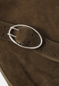 Cinturón de ante marrón oscuro con una hebilla ovalada de plata, colocado sobre una tela de ante del mismo color con costuras visibles y un borde de cremallera.