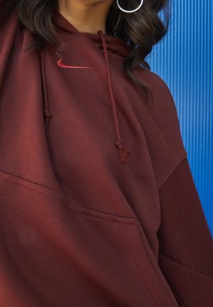 Femme portant un sweat à capuche Nike bordeaux avec cordons et boucles d'oreilles argentées en forme d'anneau, debout devant un fond texturé bleu.
