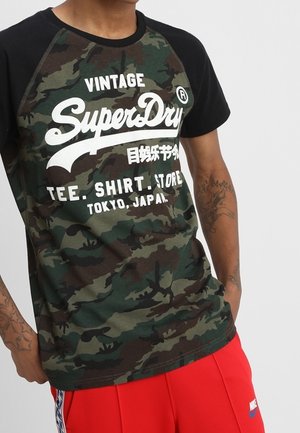 Koszulka we wzór moro z czarnym rękawem raglanowym, białym nadrukowanym logo i napisem. Wykonana z miękkiego materiału o luźnym kroju.