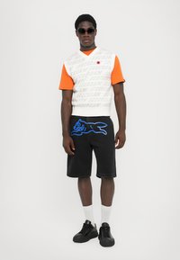 Hvid sweatervest med grå tekst, orange kortærmet skjorte, sorte denimshorts med blå hundegrafik, sorte sneakers, hvide sokker, solbriller.