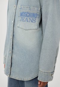 Gaiši zils džinsija krekls ar pogu priekšā, garām piedurknēm un kabatu ar uzrakstu "MOSCHINO JEANS" treknā zilā burtiem. Strukturēta auduma.