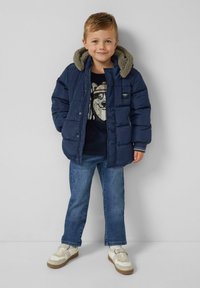 Ein kleiner Junge trägt eine marineblaue Steppjacke mit Kapuze, einen schwarzen Grafiksweater, eine blaue Jeans und weiße Sneaker, während er vor einer grauen Wand steht.