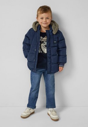 Jongens met een navy blauwe pufferjack met capuchon, een zwart graphic sweater, blauwe jeans en witte sneakers, leunend tegen een grijze muur.
