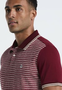 Bordeaux poloshirt met een gebreid patroon, geribde kraag met crèmekleurige accenten en een klein geborduurd logo op de linkerborst.