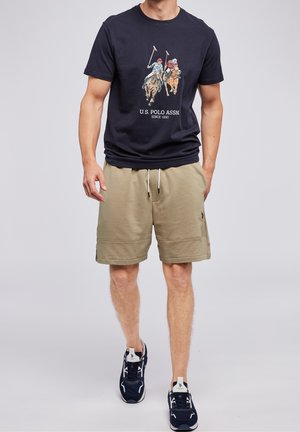 Man draagt een navy poloshirt met afbeelding van paard polo spelers, beige short met trekkoord en navy witte sneakers, lopend vooruit.
