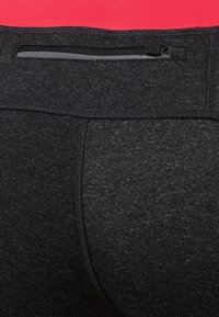 Gros plan sur un pantalon de sport noir avec une poche zippée dans la ceinture arrière et une personne portant un haut rouge.