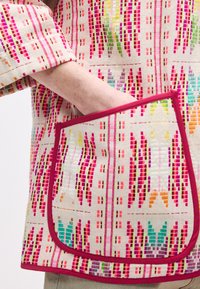 Πολύχρωμο ύφασμα με σχέδια σε ροζ, πράσινους και πορτοκαλί τόνους, με έντονο κόκκινο τελείωμα γύρω από την τσέπη και τις άκρες.