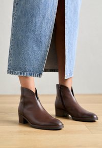 Brune skinnankelstøvler med skrå design, glidelås og lav blokkheel, parret med et denimskjørt. Teksturert overflate synlig.