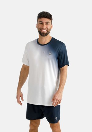 Hombre sonriente que lleva una camiseta con degradado blanco y azul marino y pantalones cortos azul marino, posando frente a un fondo liso.