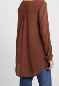 Langekleurige blouse in roestkleur met een gestructureerde stof, plooidetail aan de achterkant en een ronde onderrand. Voorzien van knoopmanchetten.