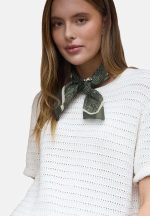 Jeune femme aux longs cheveux châtain clair portant un haut blanc à manches courtes en tricot et un foulard vert à motif noué en un nœud papillon.