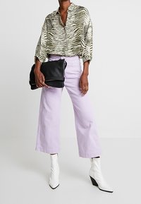Blusa a fantasia zebra in oliva e crema, abbinata a pantaloni larghi lilla e stivali con tacco bianchi alla caviglia. Borsa a tracolla nera.