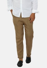 Pantaloni in lino marrone chiaro presentano un design affusolato con risvolti, abbinati a scarpe slip-on nere e una camicia bianca con bottoni.