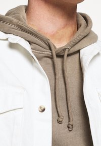 Giacca in denim bianco con chiusura a bottoni sopra un felpa con cappuccio beige, caratterizzata da polsini a costine e un cordino con estremità annodate.