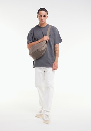 Schoudertas gemaakt van glad, beige stof met een rond ontwerp en een verstelbare strap, gedragen over een grijs T-shirt en witte broek.