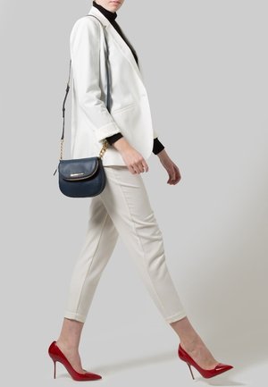 Vrouw loopt in een wit pak met een zwarte coltrui, draagt een marineblauwe crossbody tas en draagt rode hoge hakken tegen een grijze achtergrond.