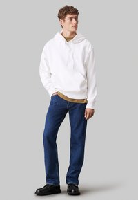 Junger Mann, der nach rechts schaut, trägt ein weißes Hoodie, ein braunes Unterhemd, blaue Jeans und schwarze Schuhe vor einem schlichten grauen Hintergrund.