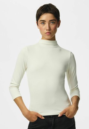 Femme aux cheveux courts et foncés portant un haut blanc à manches longues et un pantalon noir, debout les mains dans les poches devant un fond uni.