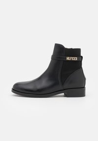 Musta nahkasaapas nilkkapituisilla ja elastisilla sivupaneeleilla sekä pienellä lohkarekorolla. Ominaisuuksiin kuuluu kultasävyinen "HILFIGER" -logotermi. Sujuva pinta.