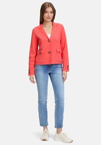 Blazer corail au design structuré, avec deux poches avant et fermeture à boutons. Associé à un jean bleu clair et des baskets blanches.