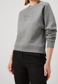 Felpa grigia con collo e polsini a coste. Logo "BOSS" in rilievo sulla parte anteriore. Texture morbida, indossata sopra pantaloni neri.