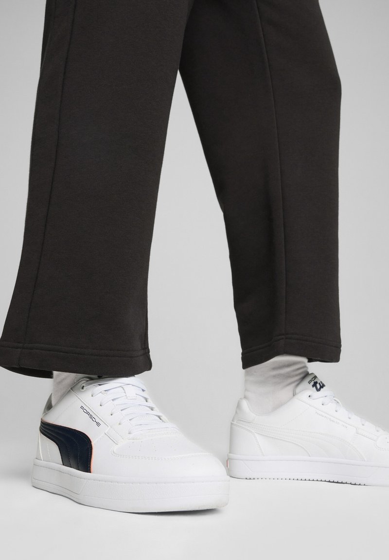 Baskets blanches Puma avec des détails de design Porsche en noir et bleu marine, portées avec un pantalon noir et des chaussettes blanches sur fond gris clair.