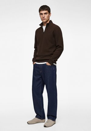 Junger Mann steht mit den Händen in den Taschen und trägt einen dunkelbraunen Pullover mit Viertel-Reißverschluss, darunter ein weißes Unterhemd, locker sitzende dunkelblaue Jeans und beige Turnschuhe.