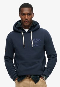 Felpa blu navy con tasca a marsupio, stringhe crema e logo. Indossata sopra una camicia a quadri verde e blu. Pantaloni beige e un orologio.
