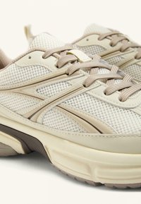 Beige Sportchuhe aus Mesh und Leder mit hellbraunen Schnürsenkeln, geschwungenen Akzenten und einer strukturierten Gummisohle.