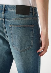 Vervaagde blauwe denim jeans met een zwart leren label aan de achterkant. Bevat twee achterzakken met contrasterende stiksels.