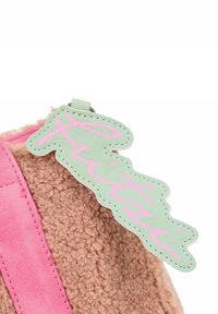 Rosa- und beigen Tasche mit weichem, strukturiertem Fell und einem grünen Lederanhänger mit pinkfarbener, geschwungener Schrift. Das Design ist kompakt und kuschelig.