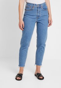 Jeans de tiro alto en azul claro con corte recto, dobladillo deshilachado y dos bolsillos delanteros, combinados con sandalias negras de deslizar con tachuelas plateadas.