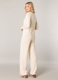 Beige Langarmoberteil, kombiniert mit weiten beigen Hosen. Glattes Material, enganliegende Passform und minimalistisches Design. Die Schuhe haben Blockabsätze.