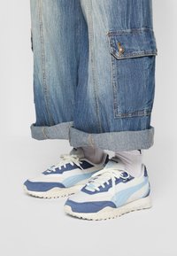 Adidași albaștri și albi din material suede și plasă; au o formă distinctivă și o talpă texturată. Purtați cu pantaloni din denim largi.