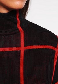 Nahaufnahme eines schwarzen gerippten Rollkragensweaters mit kräftigen roten vertikalen und horizontalen Streifen, die ein Kreuzmuster auf der Stofftextur bilden.
