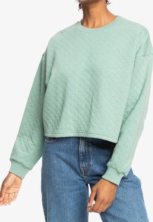 Personne portant un sweat court matelassé vert clair à manches longues associé à un jean bleu taille haute, mains détendues.