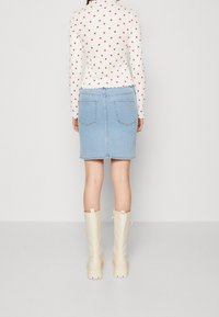 Frau, die wegschaut, trägt ein weißes Langarmoberteil mit roten Herzen, einen hellblauen Jeansrock und cremefarbene Stiefel bis zur Mitte der Waden vor einem einfarbigen Hintergrund.