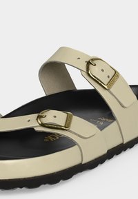 Μπεζ σανδάλι με δύο ρυθμιζόμενες χρυσές μεταλλικές αγκράφες και μαύρη διαμορφωμένη εσωτερική σόλα σε ανάγλυφη μαύρη σόλα.